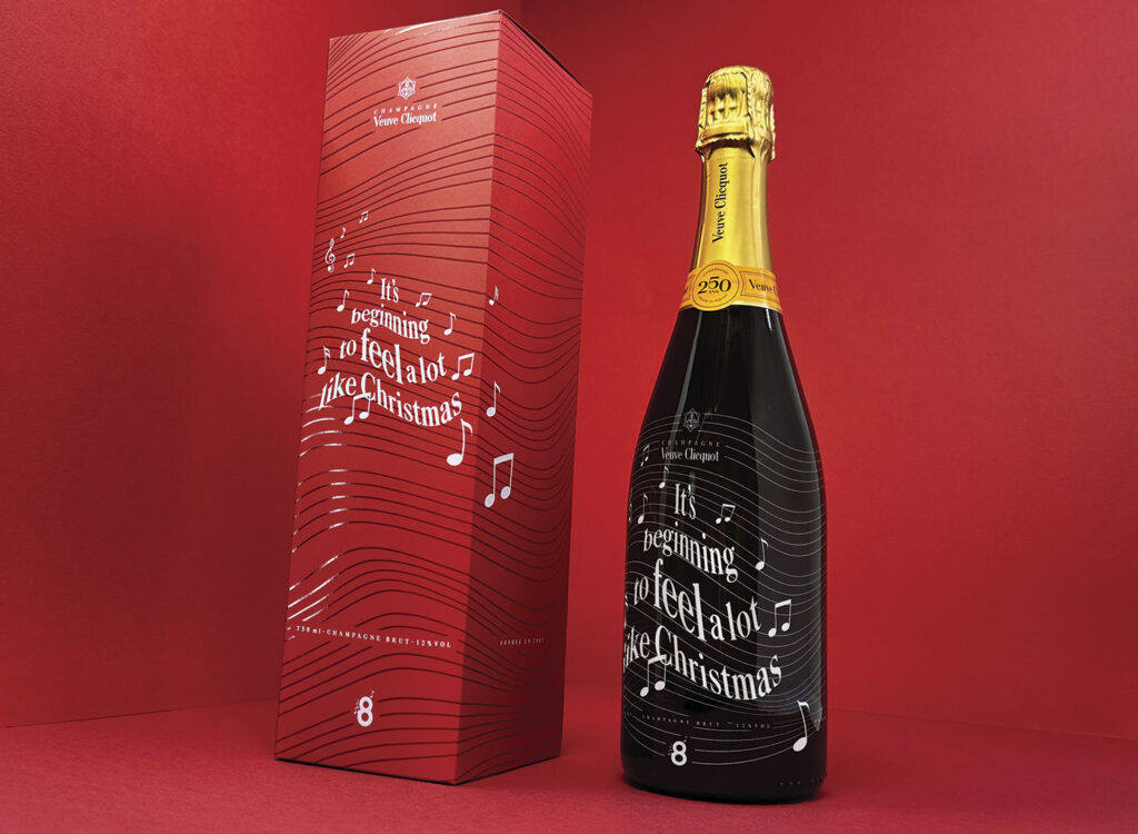 Veuve Clicquot Christmas champagne bottle with dark label beside a red gift box, both featuring white music note graphics and